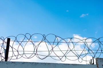 Barbed wire stretched on a concrete wall against a clear blue sky with clouds symbolizes protection...