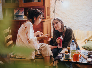 Two young women sitting indoors, involved in an engaged conversation, surrounded by cups and plants.