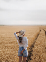 Back view to young Woman standing on field road leading across golden wheat field on a Sunny Summer Day.