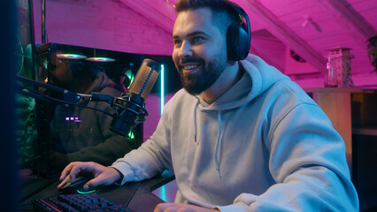 Smiling happy Caucasian addicted male gamer guy man streamer play at computer internet virtual gaming at home web battle online playing video game at dark evening with microphone and headset laughing