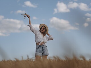 Young woman in straw hat relaxing on golden wheat field, holding brunch of ears in her hand against blue sky.