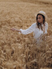 Young woman in straw hat collects bunch of ripe wheat earls in golden wheat field.