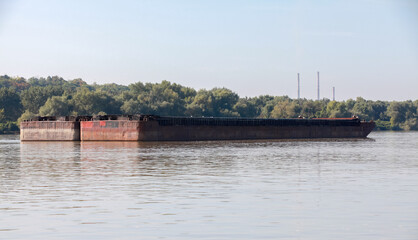 Two rusting barges float on a serene river surrounded by a lush forest
