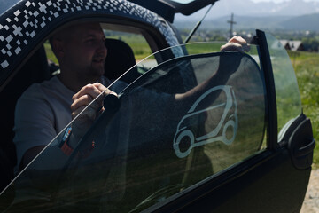 A man sitting in a small modern car, captured from the open driver's door with a scenic rural...