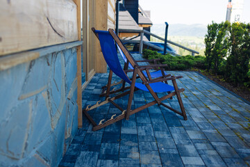 Two wooden deck chairs with blue fabric sit on a tiled terrace outside a rustic house. Surrounded by greenery and overlooking a distant mountain landscape, the scene evokes a peaceful and relaxing