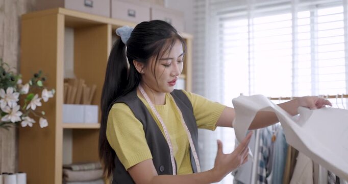 young asian female fashion designer reviewing paper pattern with focused expression measuring garment template tailoring tools fabrics placed in fashionstudio small business workspace modern setting