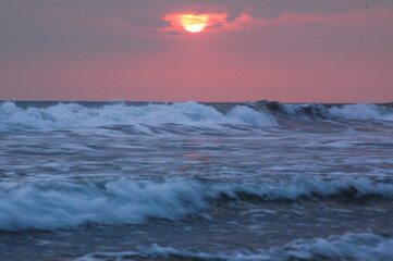 The sun sets over the ocean, casting a pink glow on the sky and waves.