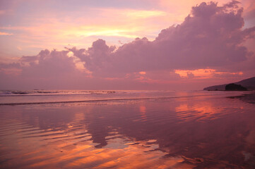 A serene beach scene unfolds at sunset, with the sky painted in hues of pink and orange, reflected on the wet sand, creating a tranquil and picturesque moment.