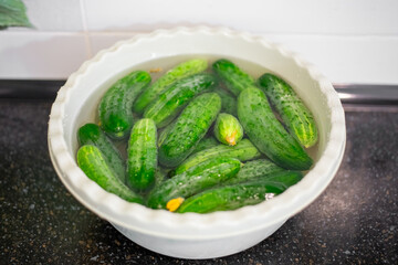 A pile of fresh green cucumbers soaking in water in a white plastic bowl on a dark surface, preparing to pickle
