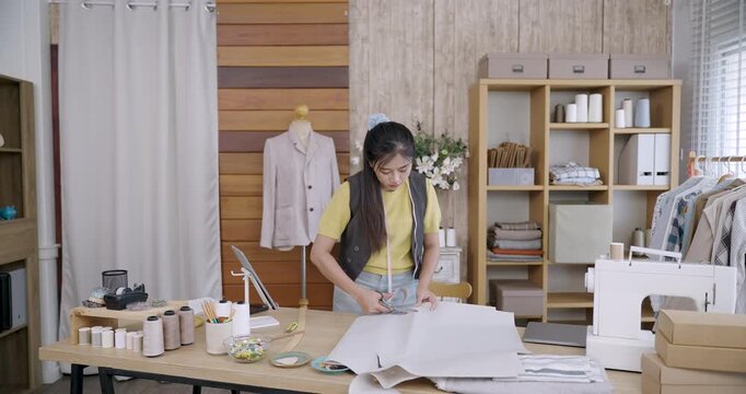 Asian female fashion designer preparing large paper pattern on table while working on garment design in tailoring studio surrounded by sewing tools and handmade clothing elements