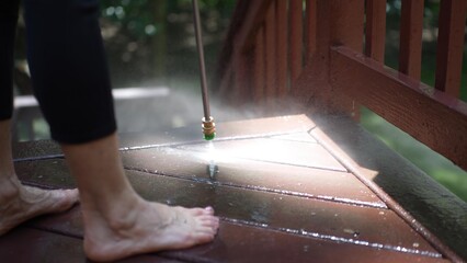 A mature woman is using a pressure washer to clean a wooden deck, removing dirt and grime while enjoying the outdoors in her backyard.