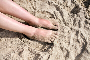 Sand-covered feet sink into sunlit beach, capturing relaxed moment. Soft texture of sand contrasts...