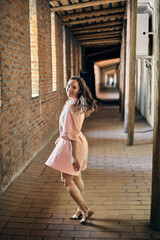 Young woman with long brown hair spins in pink dress in brick-walled corridor. Sunlight filters...