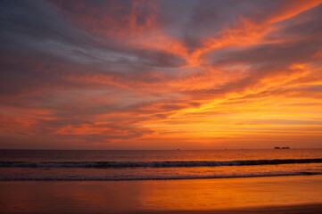 A vibrant sunset paints the sky with fiery hues of orange and red, reflecting on the tranquil ocean and sandy beach below, creating a serene and picturesque scene.