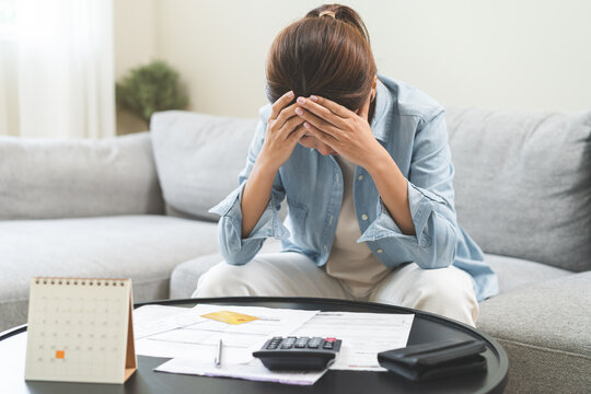 Debt and financial concept, stressed asian young woman have a headache while sitting on sofa calculate expenses, income, looking bills of credit card for prepare payment or pay bills on table at home.