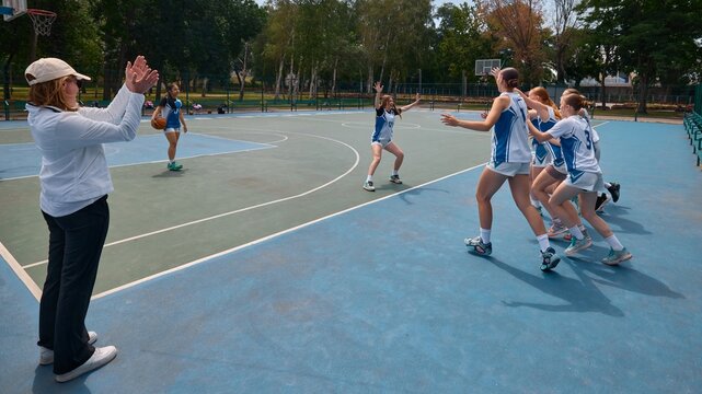 Female basketball athletes training on open court as coach claps in support. Concept of team drills, learning through repetition, female basketball development, youth growth visuals, sports promotion