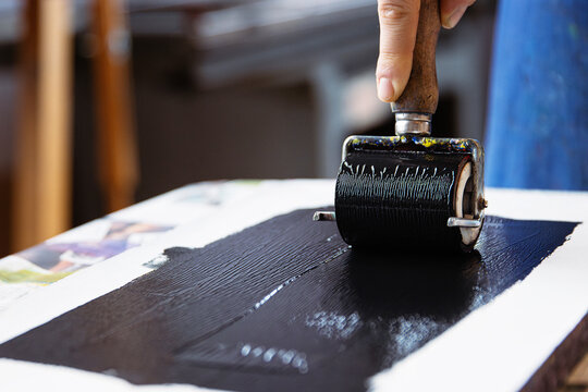 A closeup of an artist hand using a brayer to apply black ink onto a linocut surface in a linocut printmaking process.