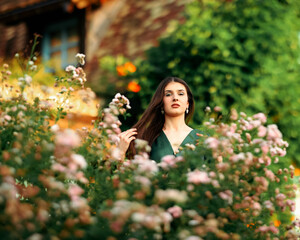Elegant woman in a green dress standing among blooming flowers in a summer garden, looking...