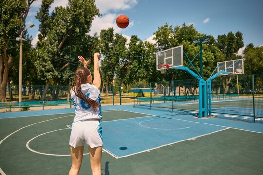 Young girl playing basketball on outdoor court beneath blue sky. Concept of shooting practice under supervision, basketball skills session, outdoor sports technique, team training branding