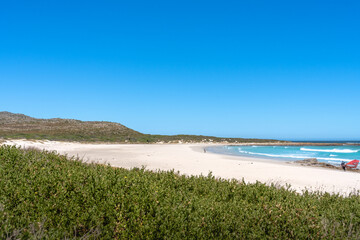 Ein breiter, unberührter Sandstrand mit türkisfarbenem Wasser und sanften Wellen, umgeben von grüner Küstenvegetation und Dünen unter klarem blauen Himmel