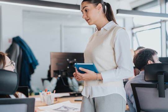 A confident businesswoman interacts with colleagues in a modern, open-plan office. The diverse workplace reflects teamwork and collaboration among multicultural employees. Ideal for concepts of