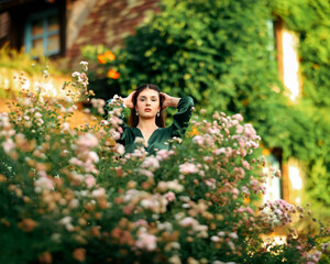 Portrait of a young woman in a green dress standing among blooming flowers in a cottage garden,...