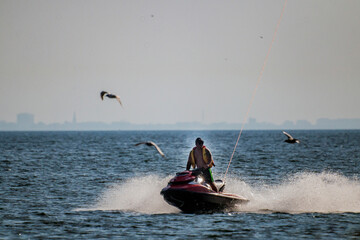 A man speeds on a red jet ski across a splashing sea as seagulls fly past