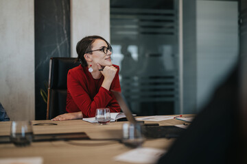 A businesswoman in a red blazer sits at a conference table, contemplating ideas and engaged in a...