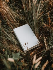 A blank, off-white book rests amidst tall grasses and wildflowers, suggesting tranquility and the outdoors.  The book's simple design contrasts with the natural textures and colors of its surroundings