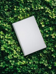 A blank, light-grey book rests on a bed of vibrant green foliage, creating a striking contrast between the man-made and natural worlds. The image's simplicity is captivating