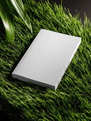 A blank, white book rests on vibrant green grass, partially obscured by a large green leaf, against a dark background.  The image suggests natural themes or environmental concepts