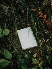 A blank, off-white book rests nestled in lush green grass and wildflowers, creating a serene, natural scene