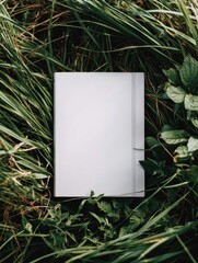 A blank, off-white book rests in a bed of vibrant green grass and dark-green foliage, creating a serene natural setting