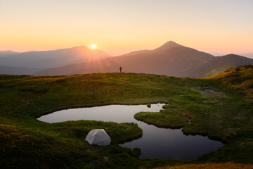 Incredible sunset on small mountain lake in summer mountains. Picturesque summer landscape with tent and silhouette of a tourist, green hills and sun rays in evening sky