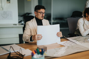 A business professional deeply examines architectural plans in a modern office environment, indicating focus and strategizing. Ideal for showcasing teamwork, project planning, and corporate analysis.