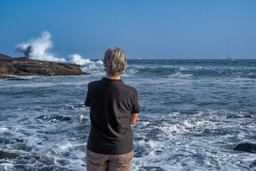 Rear view of elderly woman with short gray hair while standing by the seaside looking at the...