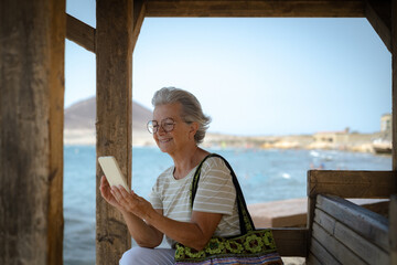 Happy caucasian senior woman sitting in the shade of the gazebo in front of the sea looking at...