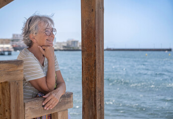 Portrait of cheerful senior woman sitting in the shade of the gazebo face the sea while looking...