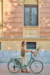 Fototapeta premium Stylish female walking next to her bicycle with groceries in the basket, promoting sustainable urban lifestyle