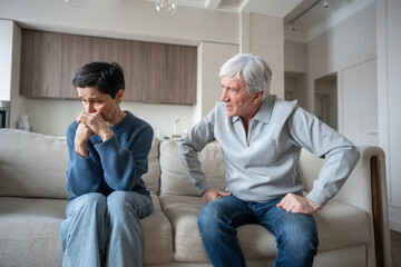Angry hysterical husband with controlling behavior toward scared wife on couch. Verbal abuse, mental blackmail. Upset mature wife showing fear and withdrawal after argument with manipulative husband