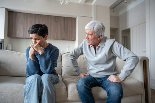 Angry hysterical husband with controlling behavior toward scared wife on couch. Verbal abuse, mental blackmail. Upset mature wife showing fear and withdrawal after argument with manipulative husband