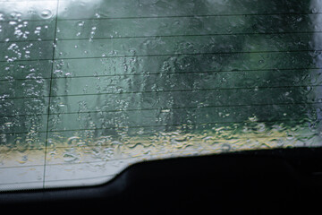 View through a car’s rear window during heavy rain, with water droplets and streaks distorting...