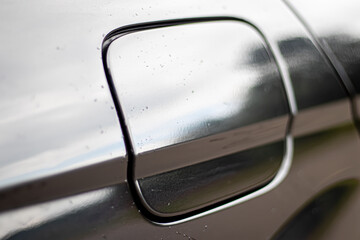 Black and white image showing a close-up of a car’s fuel tank cover, with rain droplets on the...