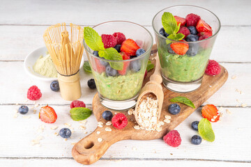 Overnight matcha oatmeal, sweet tasty oats with yogurt or non-dairy creamer, with fresh strawberries, blueberries, raspberries and mint, with ingredients on white wooden kitchen table