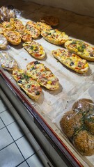 A display of various baked goods including flatbreads topped with vegetables and herbs, alongside round bread rolls. The scene is set in a bakery with a clean, tiled surface.
