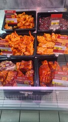 A display of various meat products in a grocery store. The trays contain chicken wings, ribs, and other marinated meats, each labeled with prices.