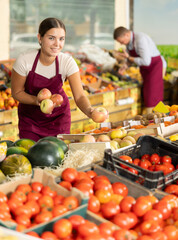 Smiling friendly young salesgirl standing at counter of organic fruit and vegetable farm store, offering ripe sweet apples