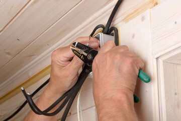 Experienced electrician carefully strips insulation from electric wire with hand tool, preparing for safe installation in wooden house.