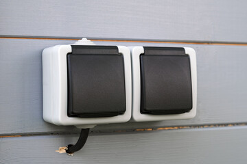 Double outdoor socket with closed black protective covers installed on gray wall offers modern waterproof power access for exterior use.