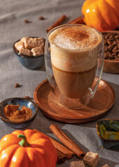 Warm pumpkin spice latte in a double glass mug surrounded by cinnamon sticks, brown sugar, and pumpkin evokes cozy autumn vibes.